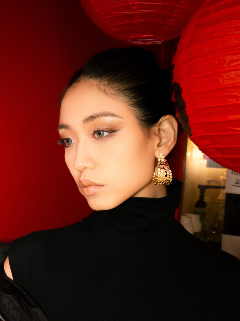Gold woven earrings against red wall and lanterns, embodying Oriental fashion elegance